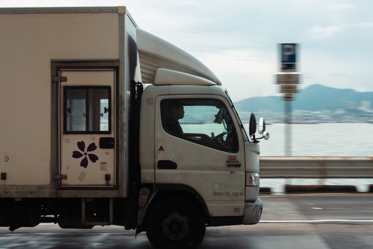 A delivery truck driving along a coastal road with a mountain backdrop on a clear day.